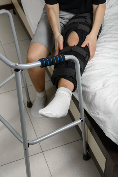 Woman wearing adjustable knee brace after leg fracture sitting on bed trying to get up with walker. Recovery and rehabilitation after surgery and installation of plates - Fotografie, Obrázek