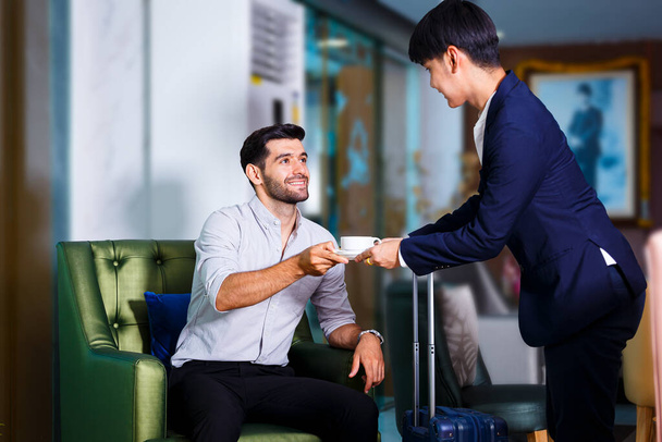 Jonge zakenman in shirt met koffer zittend op de bank in de lobby van het hotel, Receptionist man die hete koffie geeft aan jonge gasten man, ze glimlachen samen - Foto, afbeelding
