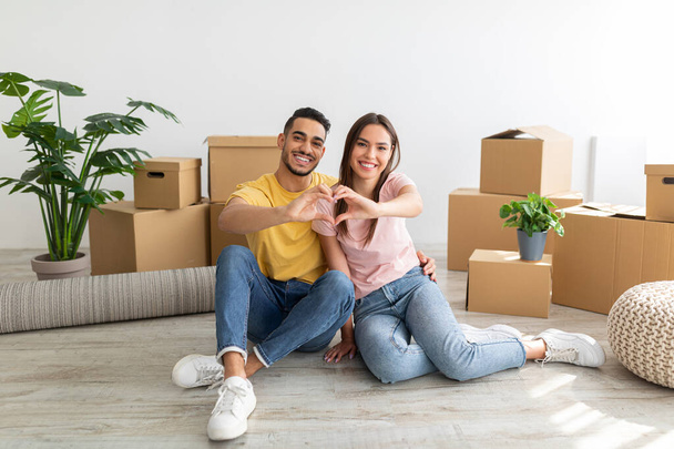 Loving young international couple making heart with their hands, sitting on floor among boxes, moving to new house - Zdjęcie, obraz