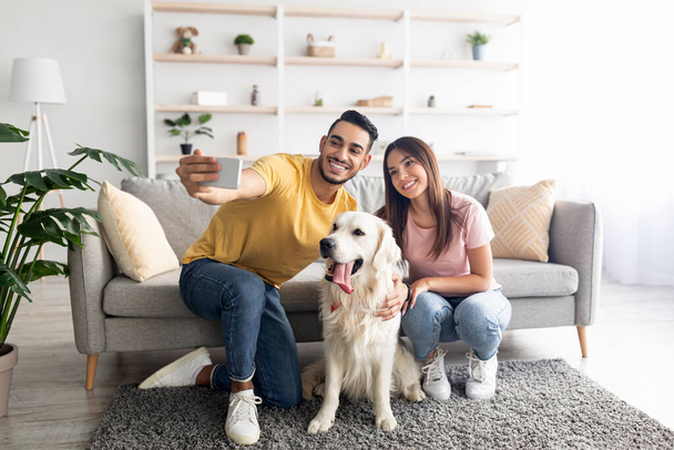 Positive young diverse couple taking selfie with their dog while sitting on floor at home, full length - Valokuva, kuva