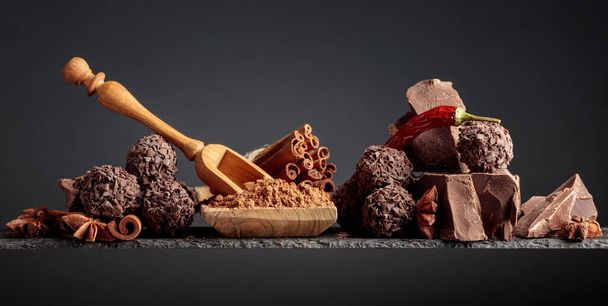 Chocolate truffles, broken pieces of black chocolate, and spices. Chocolate, cocoa powder, cinnamon sticks, red pepper, and anise on a dark background.  - Zdjęcie, obraz