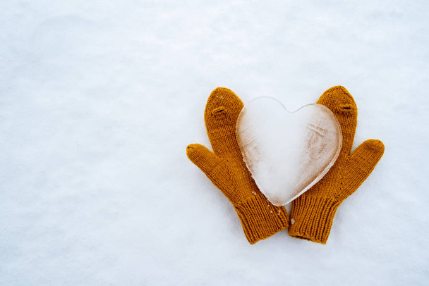 Corazón símbolo del amor, hecho de hielo, en cálidos manoplas de punto amarillo, en la nieve. Día de San Valentín. Copiar espacio. Conceptual. Vista superior.  - Foto, imagen