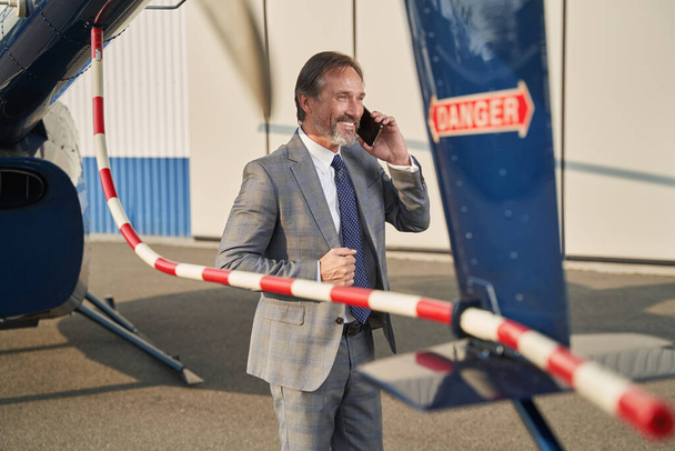 Joyous businessman talking on smartphone beside rotorcraft - Foto, afbeelding