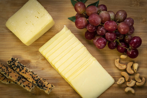 Sliced cheddar cheese block on wood board with grapes, cashew nuts, bread sticks and honey - Valokuva, kuva
