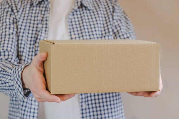 Courier in a check shirt holds out a sealed cardboard box. Delivery of online orders. The delivery clerk brought the packaged order. Space for text. - Photo, image