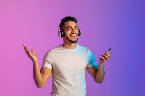 Cool Arab man listening to popular song in headphones, checking new mobile music app on phone, relaxing in neon light - Valokuva, kuva