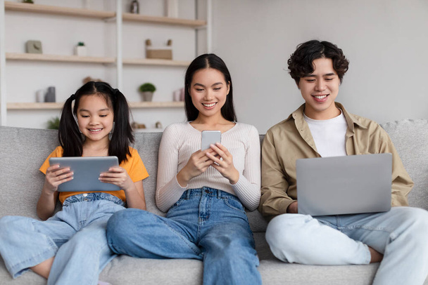 Smiling teen child and young asian mother and father surfing in internet, chatting on gadgets, play game - Фото, изображение