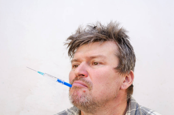 Portrait of a weirdo holding a blue syringe in his mouth. Unshaven man holding a disposable syringe full of liquid. Adult male with ruffled hair.  Indoors. Selective focus. - Photo, Image