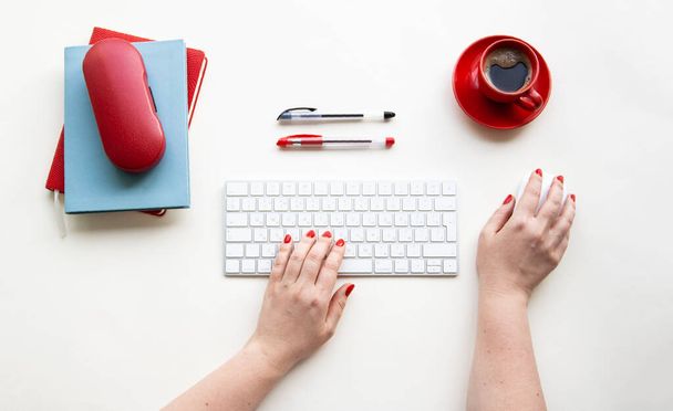 Руки жінки на бездротовій клавіатурі та миші з чашкою кави, ручок, блокнотів, футлярів окулярів на білому столі
 - Фото, зображення