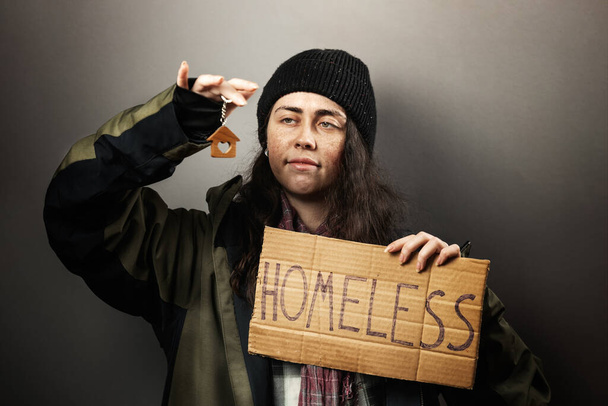 Μια ζητιάνα χαμογελαστή γυναίκα κρατά μια χάρτινη πινακίδα με την επιγραφή άστεγη και κοιτάζει ένα μπρελόκ σε μορφή σπιτιού. Η έννοια της βοήθειας περιπλανώμενους. - Φωτογραφία, εικόνα
