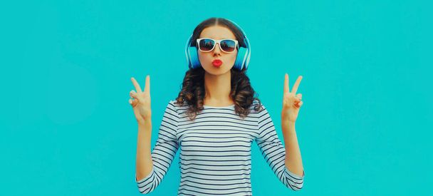 Portrait of stylish young woman in headphones listening to music blowing her lips sends sweet air kiss on blue background - Zdjęcie, obraz