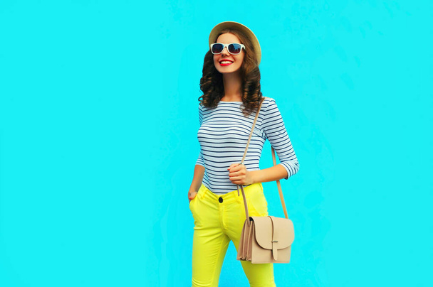 Portrait of happy smiling stylish young woman posing wearing a handbag, striped t-shirt and summer straw round hat on blue background - Foto, Bild