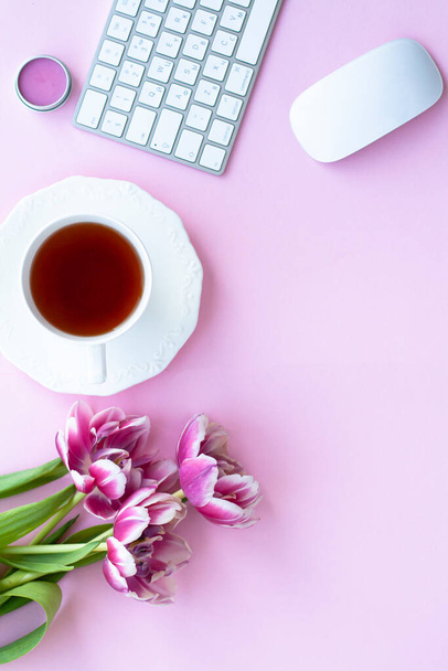 Tasse schwarzer Tee mit Untertasse, drei rosa Tulpen, kabellose weiße Tastatur und Maus, Lippenbalsam auf rosa Kopierfläche - Foto, Bild