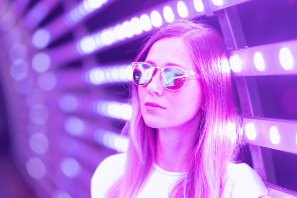 Teen hipster girl in stylish glasses standing on purple tunnel with neon light wall background, female teenager fashion model pretty young woman looking at night club city light glow - Foto, immagini