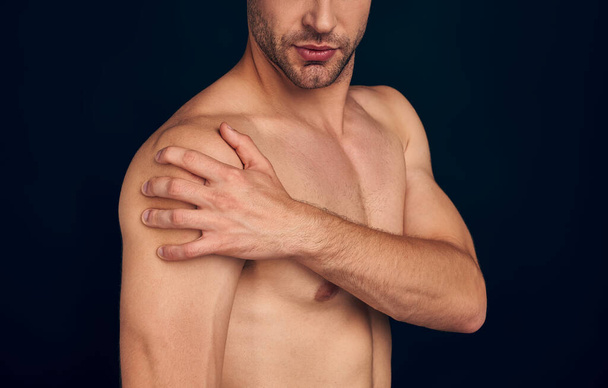 Handsome young bearded man isolated. Portrait of naked muscular man is standing on dark blue background. Man holding his shoulder. Experiencing shoulder pain. - Foto, immagini