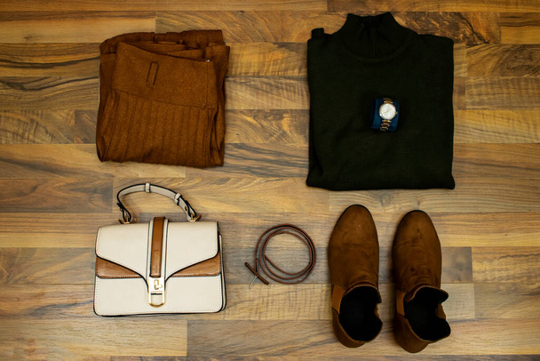 Un conjunto de ropa y accesorios de moda. suéter verde y falda corrugada, bolso de mano, botas de tobillo y cinturón de cuero marrón en la mesa de madera, disposición plana. Estar en tendencia - Foto, imagen