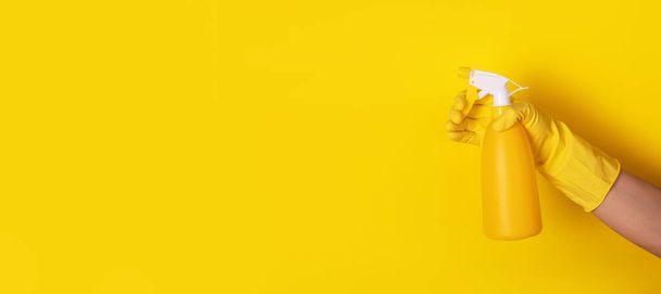 hand in een handschoen met een spray fles over gele achtergrond, netheid concept, panoramische lay-out - Foto, afbeelding
