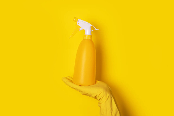 mano en un guante sosteniendo una botella de spray sobre fondo amarillo, concepto de limpieza - Foto, Imagen