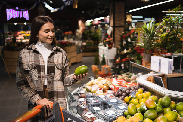 Happy Young Woman Choosing Ripe Mango in Grocery Store - Fotografie, Obrázek