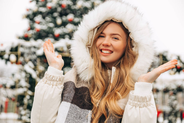 Festive Christmas fair, winter holidays concept: happy smiling woman in a scarf, catching snowflakes, posing in the city near a festive decorated Christmas tree, New Year - Fotó, kép