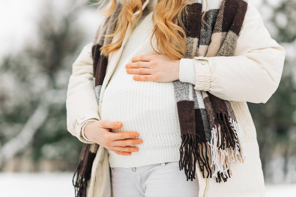 pregnant woman in warm winter clothes standing outside on a snowy winter day, with hands on her stomach on the background of nature. Concept of pregnancy, motherhood, expecting the birth of a child - Foto, afbeelding