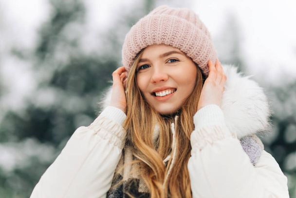 Image of a happy young woman, in a warm knitted scarf and hat looking at the camera, woman posing in the street in a snowy winter park, Winter vacation concept, winter clothes - 写真・画像