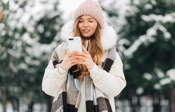 beautiful woman in a knitted hat and a warm coat and scarf walks in the winter park and uses a mobile phone. Woman in white knitted sweater. Girl using phone - Zdjęcie, obraz