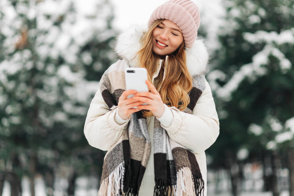 beautiful woman in a knitted hat and a warm coat and scarf walks in the winter park and uses a mobile phone. Woman in white knitted sweater. Girl using phone - Photo, Image