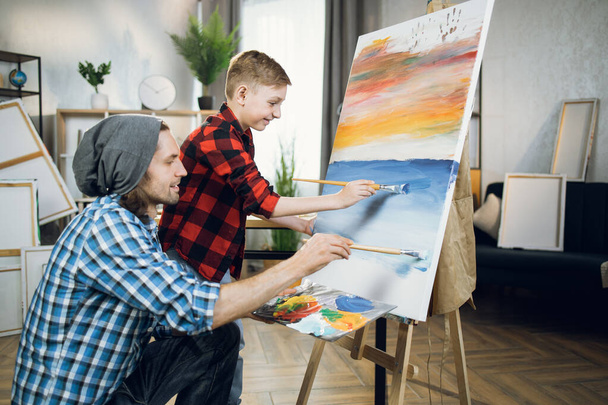 Male teacher drawing with little boy on canvas during lesson - Foto, imagen
