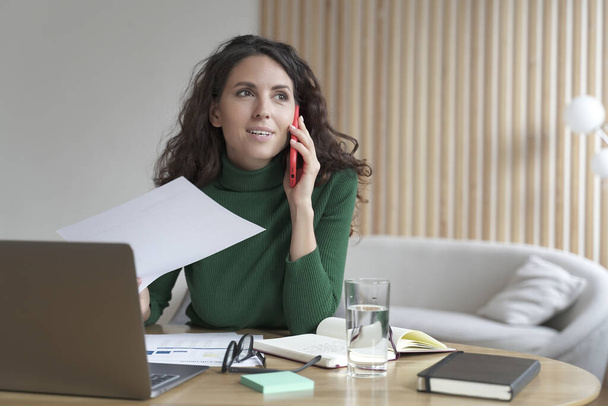 Glimlachende Italiaanse zakenvrouw zitten aan tafel bellen op haar werkplek tijdens het werken op laptop - Foto, afbeelding
