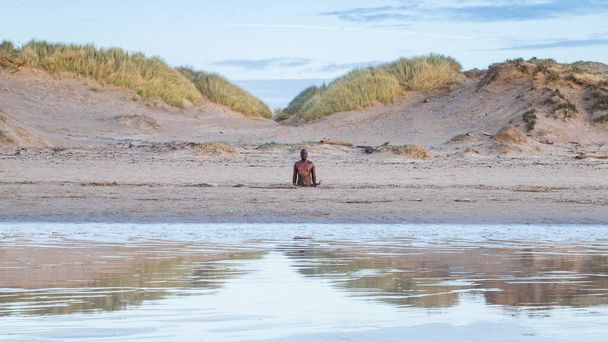 クロスビービーチの砂丘の間で見たアイアンマン。彼は、アントニー・ゴームリーによって作成されたビーチで99人の他の人物とともに、 2022年1月の夕暮れ前の湿った砂の中に反映されて見られました。. - 写真・画像