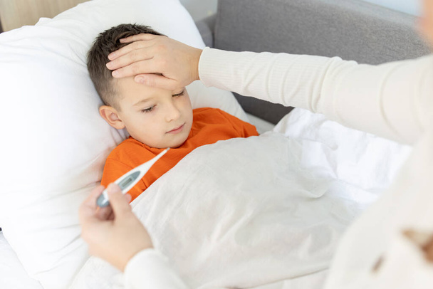 Una madre mide la temperatura de su hijo enfermo. Un niño enfermo con una alta temperatura yace en la cama y la madre sostiene un termómetro. - Foto, imagen