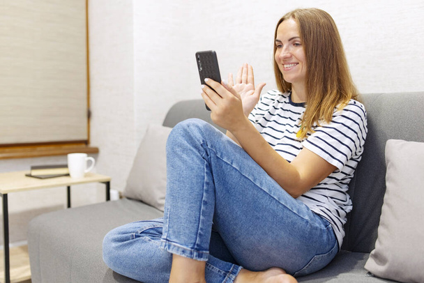 Cheerful young woman is using smartphone for video call, gesturing hi to friend or parent. Smiling young caucasian woman sits relax on couch in living room talks on video call. - Фото, изображение