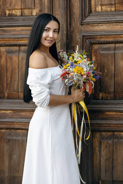 A lovely brunette girl in a stylish white dress holds a bouquet of wild dry wildflowers against the background of an old brown door. Vertical portrait of chic model holding flowers in vintage style - Photo, Image