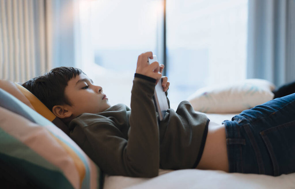 Happy Kid lying in bed holding tablet watching cartoon and chatting with friends on digital pad, Cute young boy playing games online on internet, Child relaxing at home with family - Foto, afbeelding