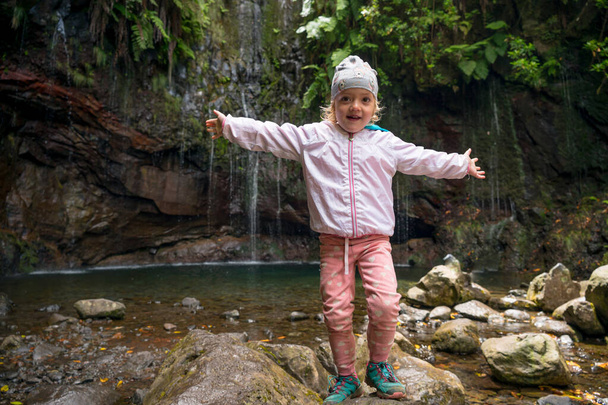 Traveler girl wearing sport jacket and raising hands at waterfall. Travel lifestyle. View from back. - Foto, afbeelding