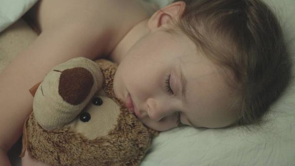 little baby sleeps at night in crib an embrace with teddy bear, girl daughter dreams while lying bed with her eyes closed, child with friend with doll bear rest together, kid face and toys close-up - 写真・画像