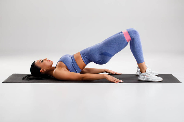 Mujer fitness haciendo ejercicios de puente de glúteos con banda de resistencia sobre fondo gris. Chica atlética haciendo ejercicio - Foto, Imagen