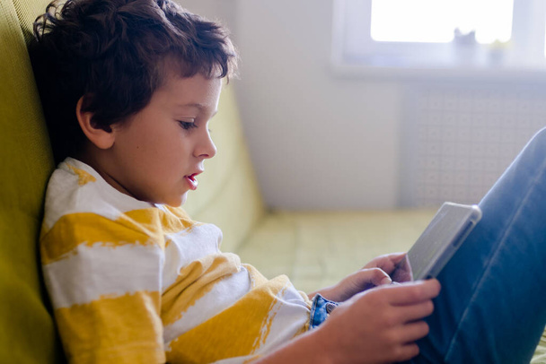 jongen in geel en wit t-shirt speelt enthousiast een spel op een tablet - Foto, afbeelding