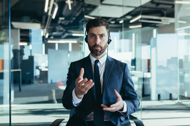 The employee makes a video call, looks at the camera and explains the task, serious gestures with his hands works in a modern office, uses a headset - Fotó, kép