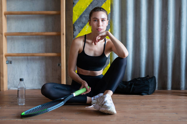 Portrait of female tennis player with racket sitting on gym floor - Zdjęcie, obraz