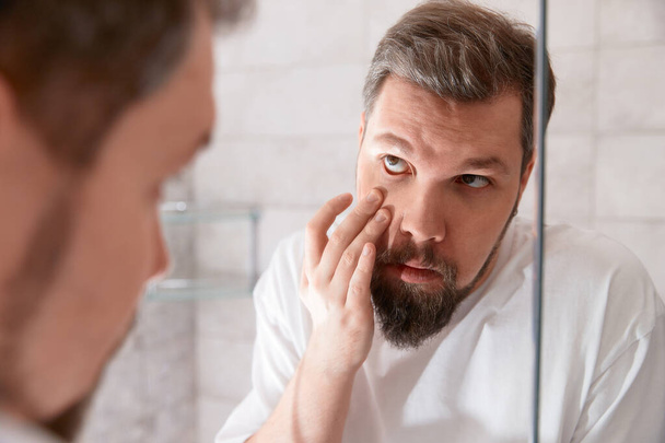 Portrait of man in white tshirt looking at the eye in the mirror. Healthcare concept. - Photo, image