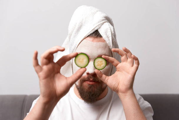 Portrait of funny happy man in white tshirt applying cucumber slices over clay mask on his face. Self care morning spa procedure - Fotoğraf, Görsel