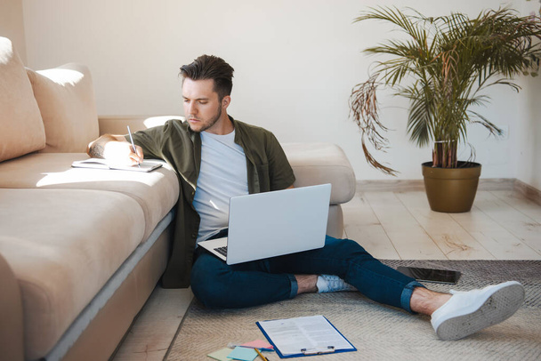 Μπροστά όψη ενός άντρα που γράφει σε χαρτί ενώ κάθεται στο πάτωμα με το laptop. Τεχνολογία Διαδικτύου. Ηλεκτρονική εκπαίδευση. - Φωτογραφία, εικόνα