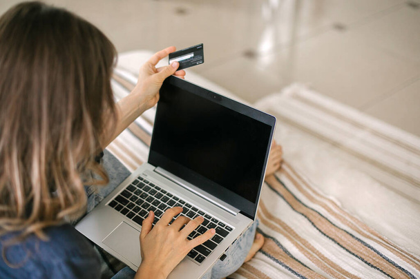 Cropped shot of asian woman holding credit card while doing shopping online on modern laptop or making money transfer through internet while sitting on bed at home, selective focus on female - Zdjęcie, obraz