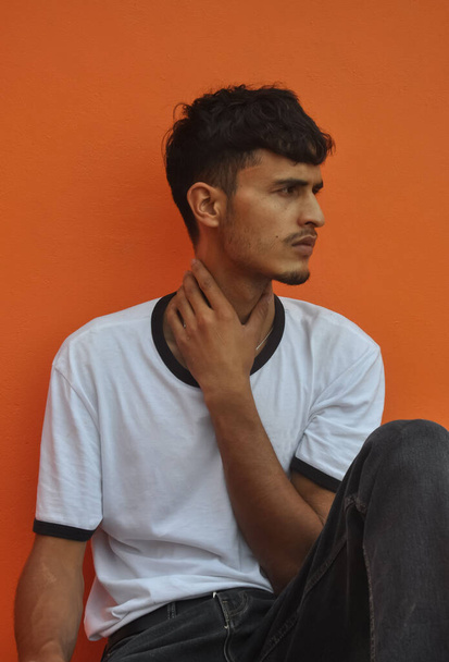 Dobře vypadající indický mladý chlap dívá bokem, zatímco sedí venku pózovat proti oranžové zdi pozadí s kopírovacím prostorem  - Fotografie, Obrázek