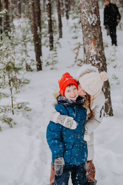 mom kisses her son in the winter forest. Mom walks with her son in a snowfall in the forest. Happy winter holidays with snow. Winter walks in the forest. Mom's love for her son. - Photo, Image
