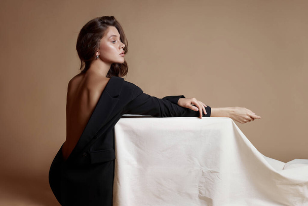 黒いジャケットのブロンドの巻き毛が白いテーブルに座っている。完璧なスリムなボディナチュラルメイク。長い髪、天然化粧品 - 写真・画像
