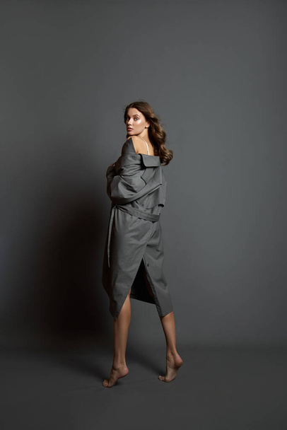 Beautiful woman in a beige bodysuit and a long gray raincoat poses standing on a gray background. Long beautiful legs - Photo, image