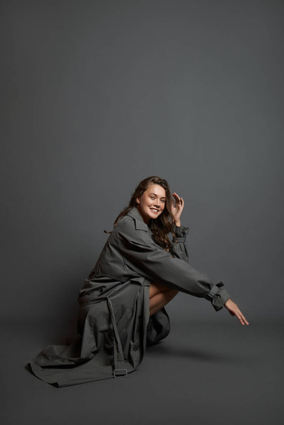 Beautiful woman in beige bodysuit and long gray raincoat poses sitting on the floor on a gray background. Long beautiful legs of a fashion model - Fotó, kép